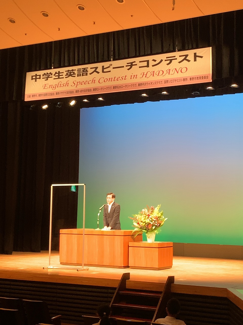 「中学生英語スピーチコンテスト」の横断幕が掲げられたステージ上で、スーツ姿の市長が華やかな生け花が飾られている演台でスピーチをしている写真