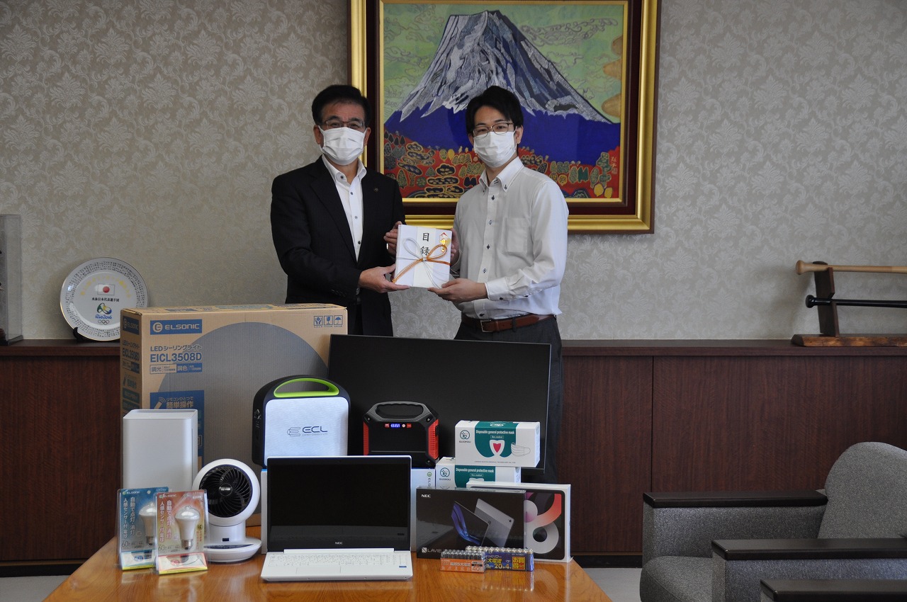 富士山の絵画が飾られた部屋で、中央のテーブルに置かれた家電製品の寄贈品を前にして、マスクを着用した市長と男性がのし袋を持って立っている写真