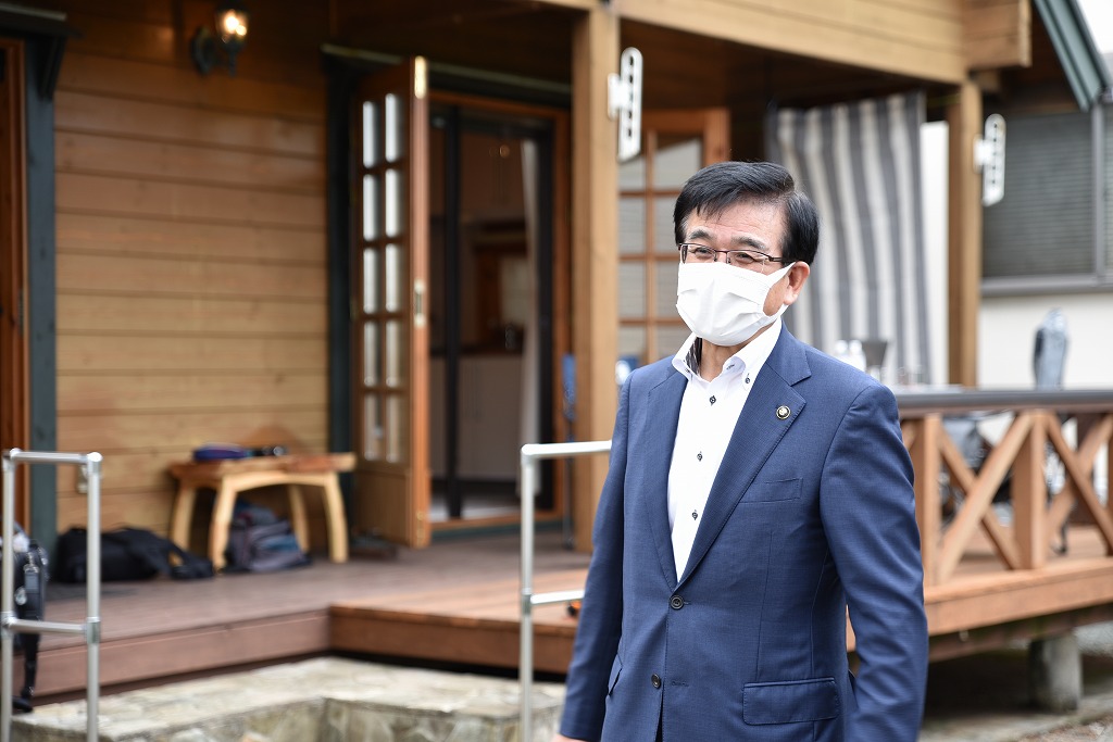 ウッドデッキとログハウス風の木造の建物前に、マスクを着用したスーツ姿の市長が立っている写真