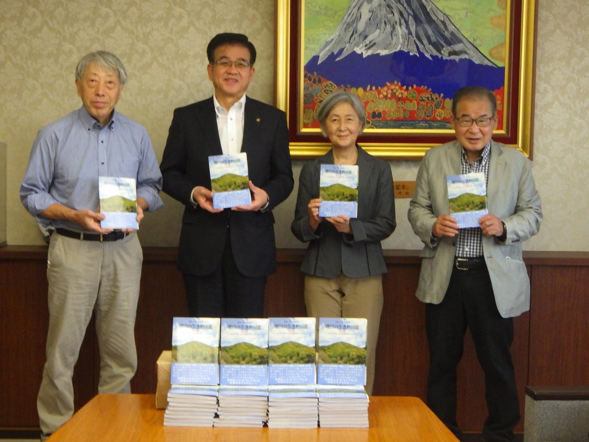 富士山の絵画が飾られた部屋で、市長と3人の男女が、たくさんの書籍を前に、それぞれ本を持って記念撮影をしている写真