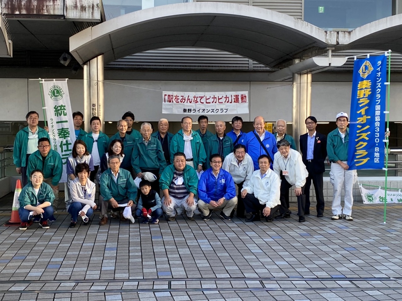 緑色のジャンパーや上着を着用した多くの人々が、駅前で「統一奉仕デー」ののぼりや「駅をみんなでピカピカ運動」と書かれた横断幕の前で集合写真を撮っている写真