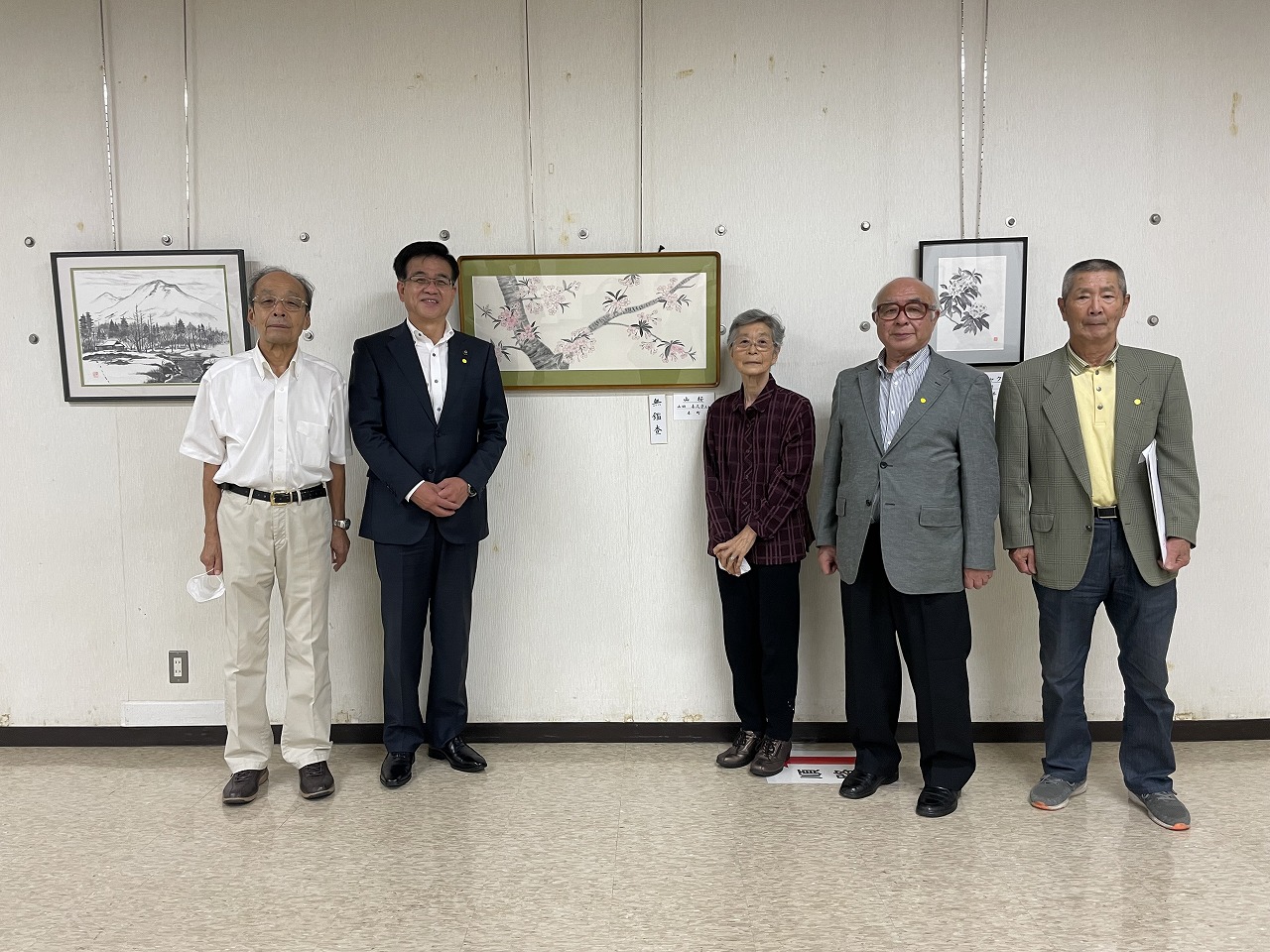 白壁に絵画が飾られた展示室で、桜が描かれた絵を挟んで左側に高橋市長と男性、右側に年配の女性と男性2名が並んで記念撮影をしている様子の写真