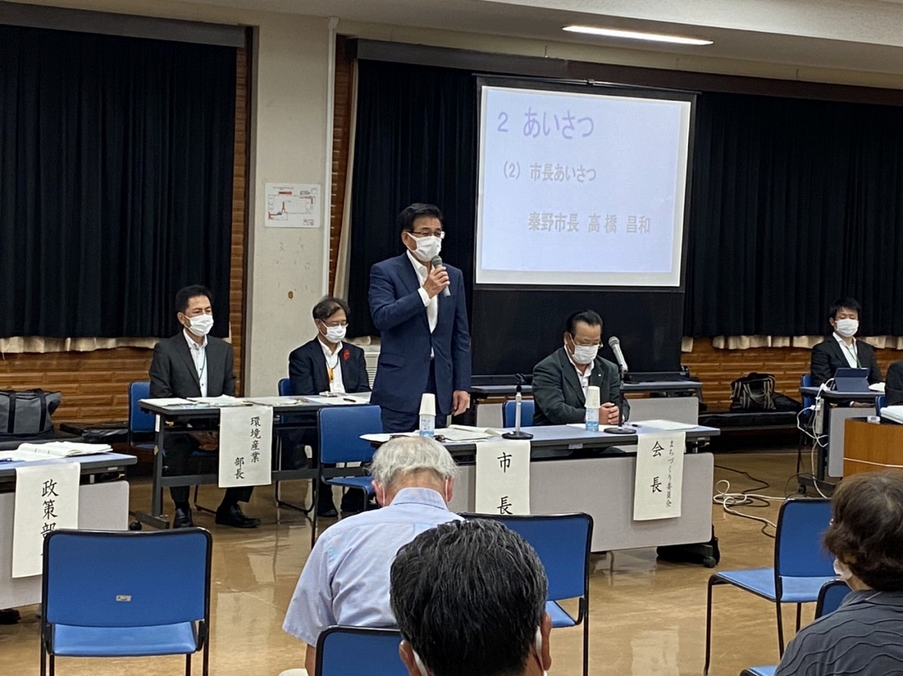 「市長あいさつ」「秦野市長 高橋 昌和」の文字が投影されているスクリーンが設置された室内で、高橋市長がマイクを持って挨拶をしている様子の写真