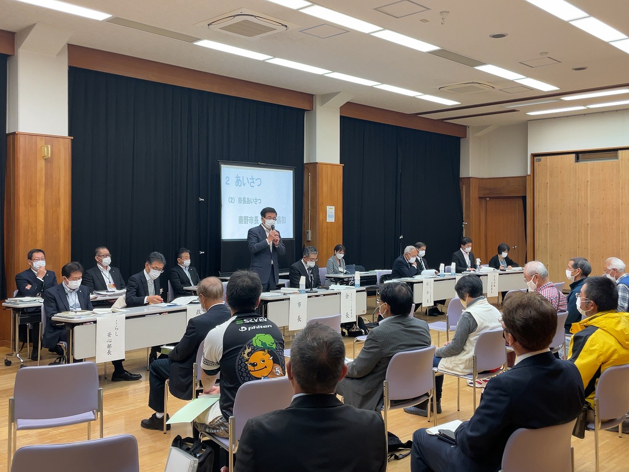 室内の前方に設置されたテーブル席に座る人々と向かい合わせに並べられた椅子に座る参加者たちがいる中、設置されたスクリーンの前で高橋市長がマイクを持って挨拶をしている写真