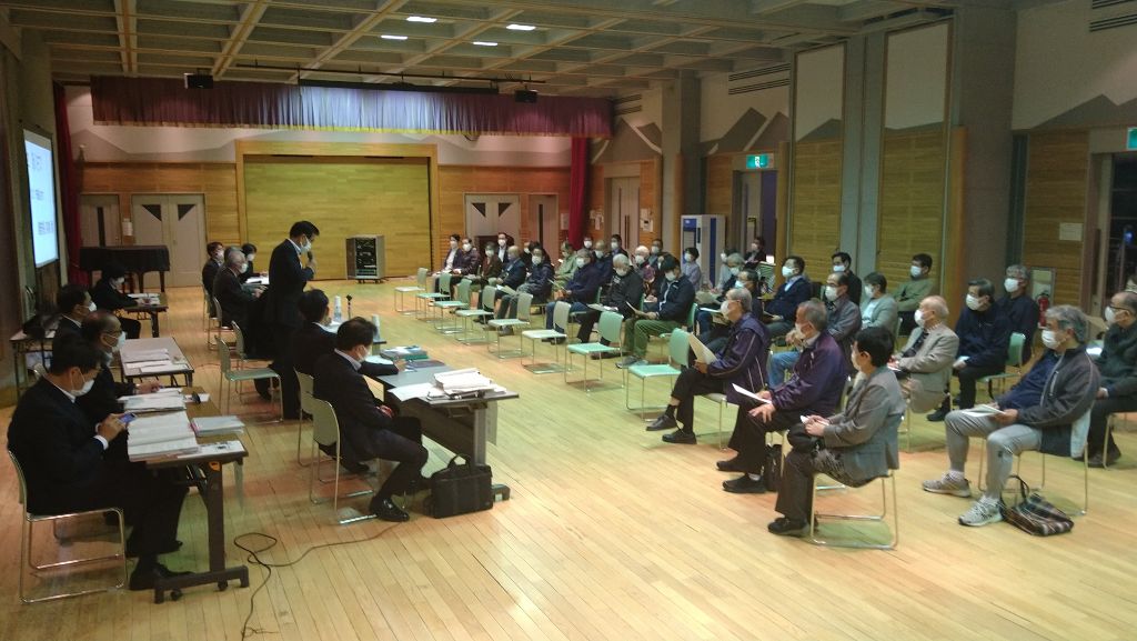 講堂のような広い部屋で、マスクを着用した多くの人々が、机に座っているスーツ姿の男性たちの方を向いて椅子に座っている懇談会の様子を撮影した写真