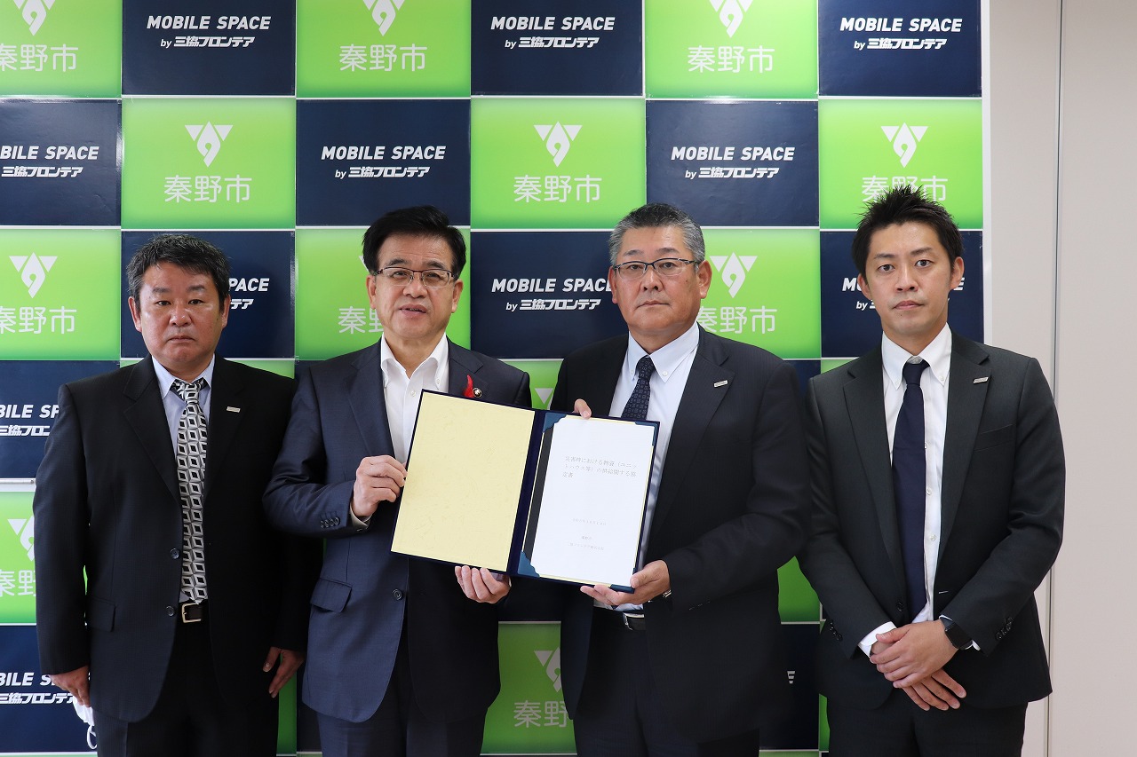 バックパネルを背景に、スーツ姿の高橋市長と三協フロンテア株式会社の関係者の男性3名がカメラに向かって立ち、高橋市長と男性が協定書を持って写真に写る様子を捉えた写真
