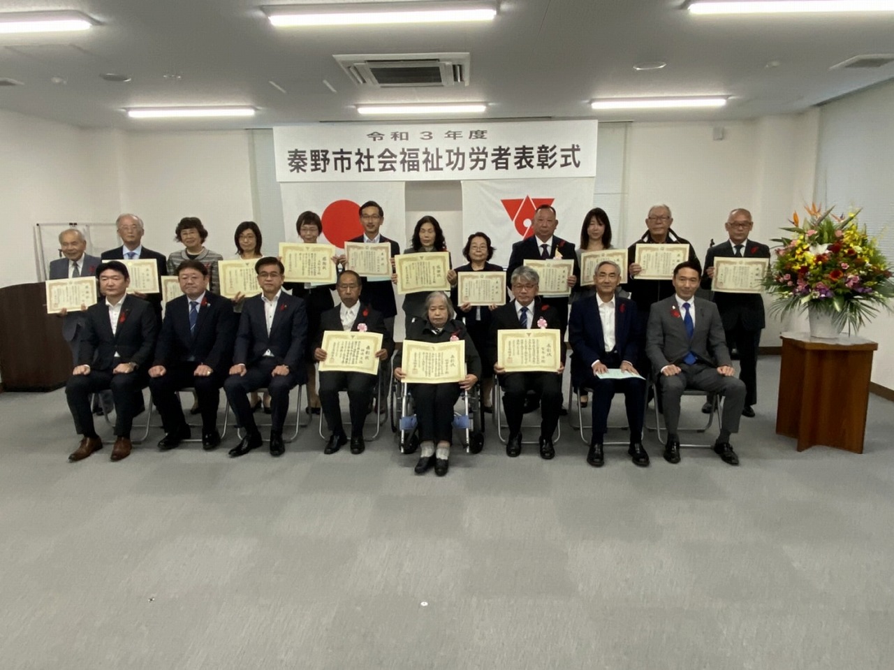椅子に座る人々と後列に立つ人々が、秦野市社会福祉功労者表彰式の横断幕を背景に表彰状を手に持って笑顔で記念撮影に応じる様子を捉えた写真