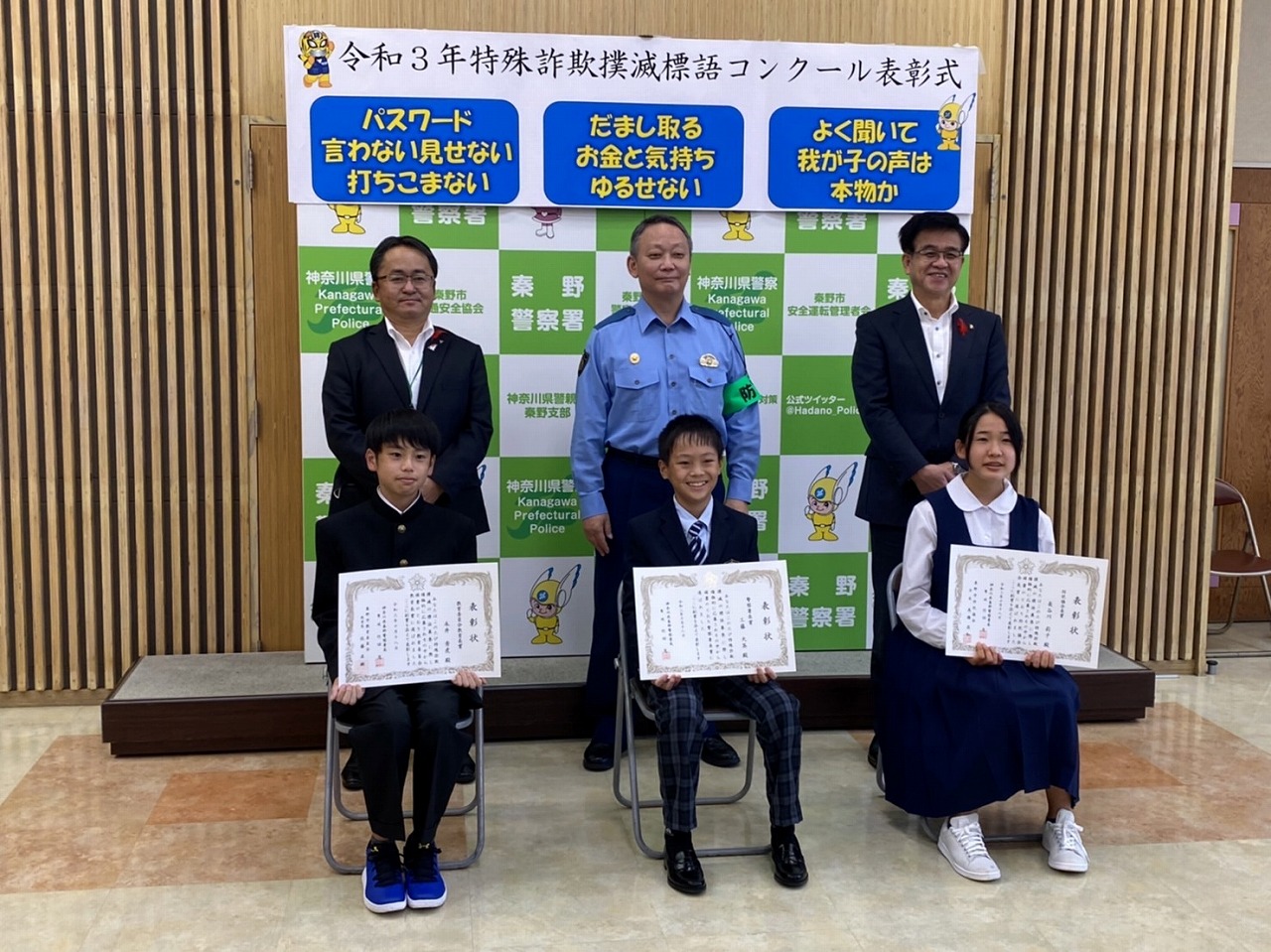 和やかな雰囲気の中、秦野警察署の警察官と高橋市長、スーツ姿の男性が、表彰状を授与された3名の学生たちとともに記念撮影をしている様子を写した写真