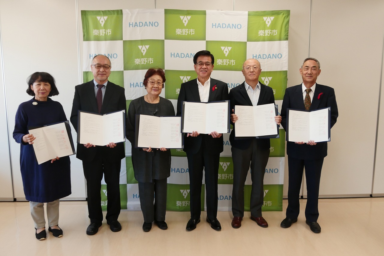 秦野市のバックパネルの前で、高橋市長を中心に5名の男女がそれぞれ協定書を開いて持ち記念撮影をしている写真
