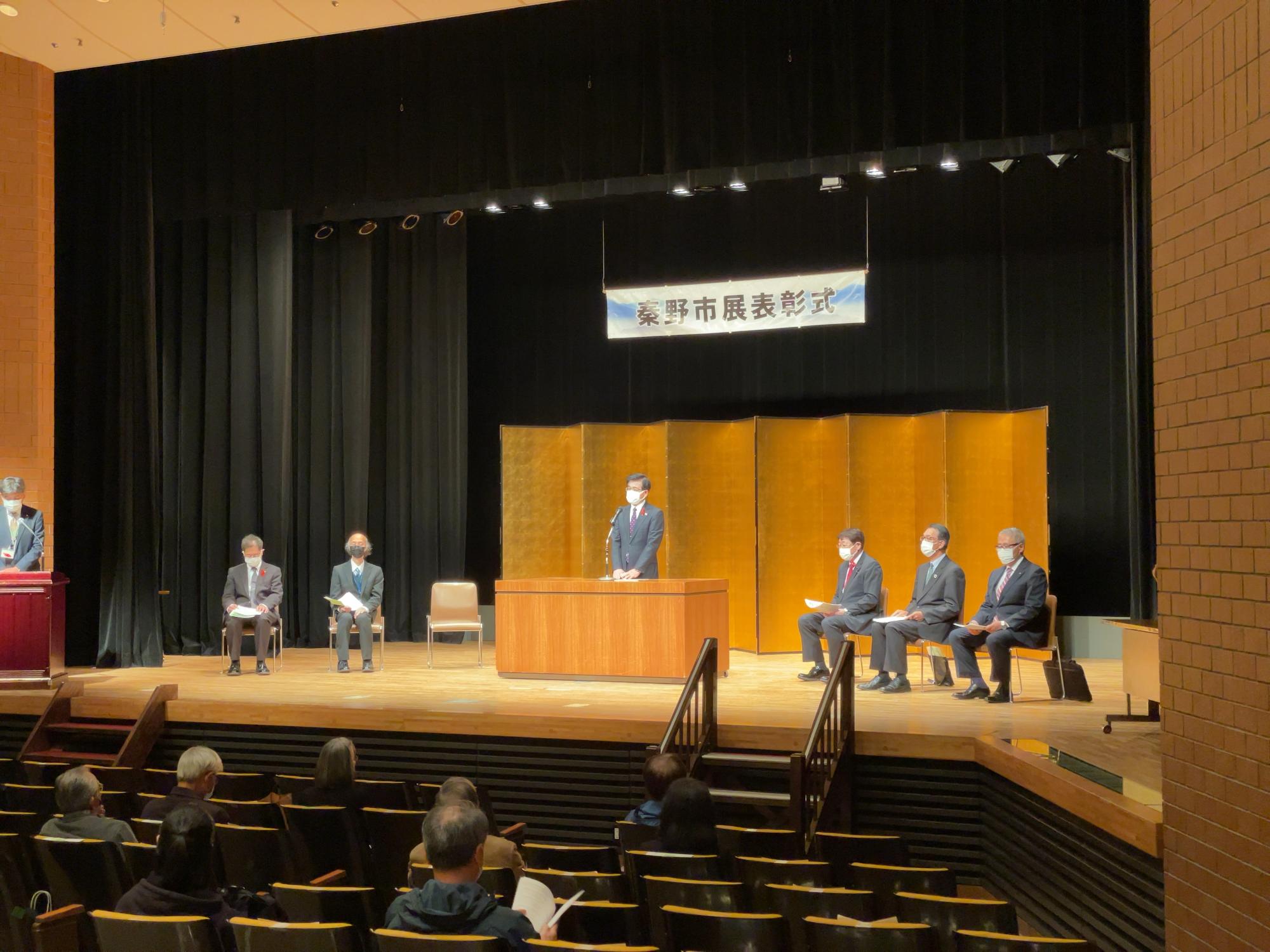 金色の屏風を背景に「秦野市展表彰式」の横断幕が掲げられたステージで男性5人が座り高橋市長が壇上に立つ写真