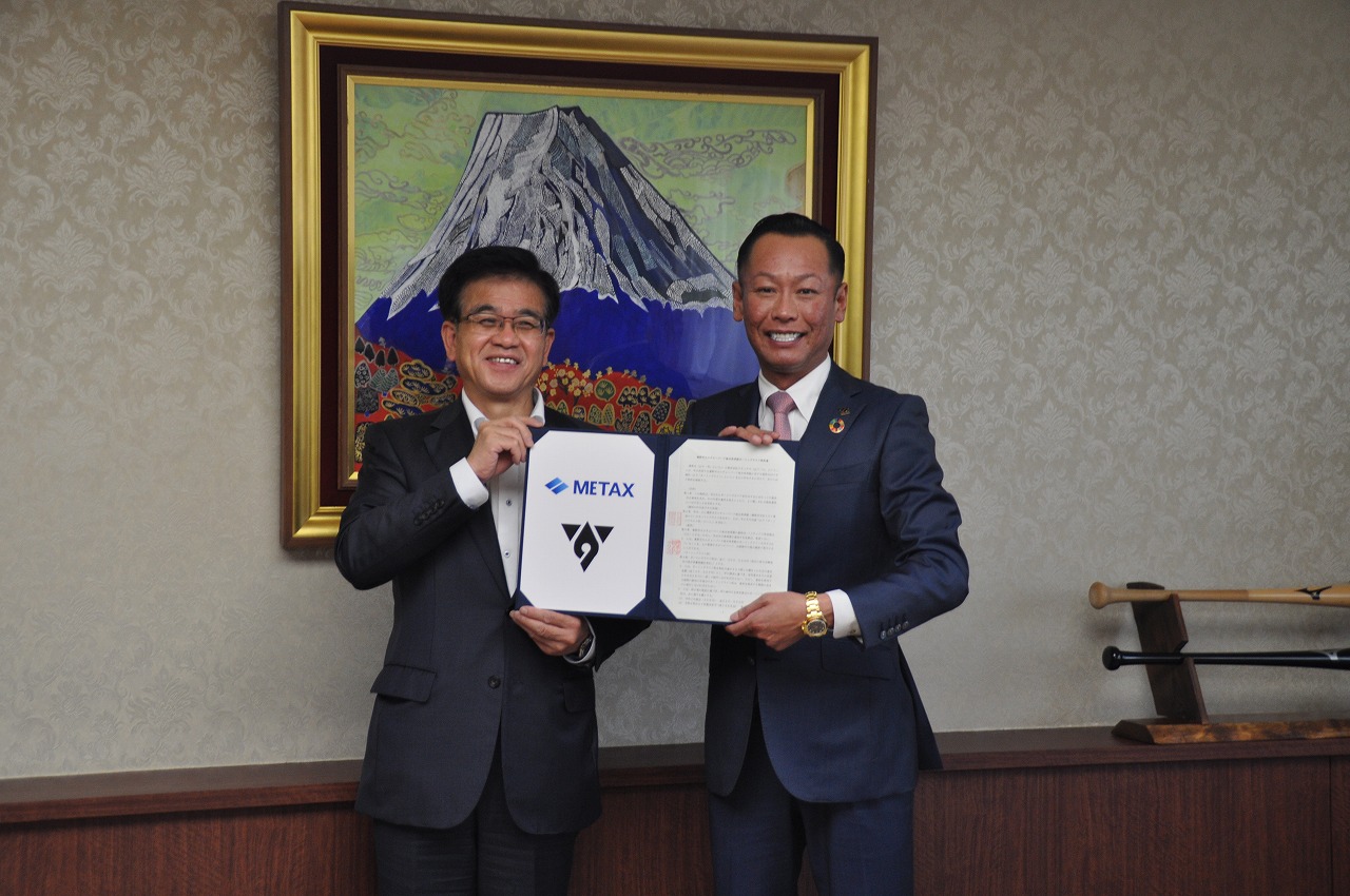 スーツ姿の高橋市長と男性が満面の笑みを浮かべ、METAXのロゴが入った協定書を持ってカメラ目線で並び立つ様子を写した写真