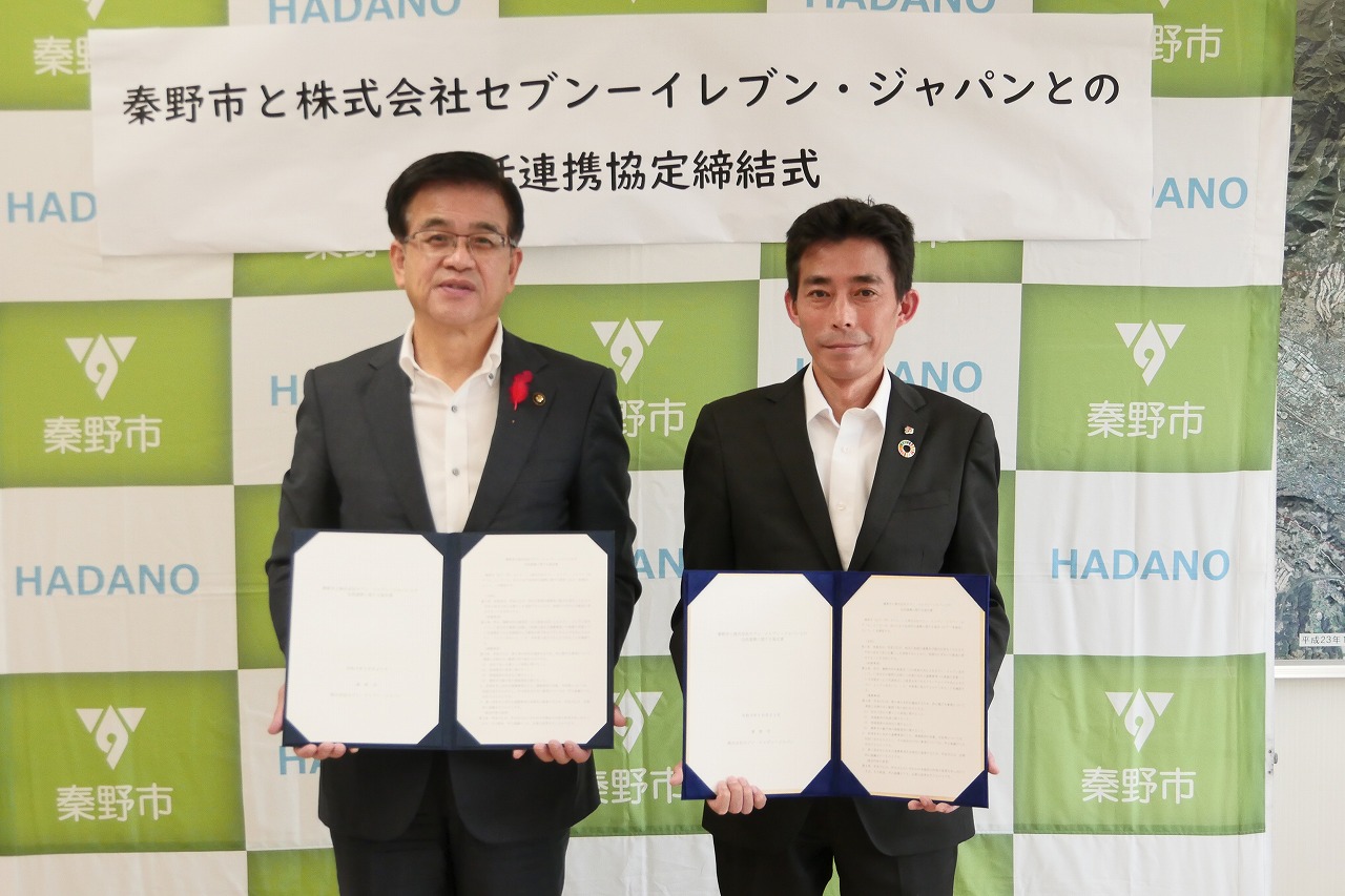 秦野市のバックパネルの前で、高橋市長と株式会社セブン イレブン・ジャパンの男性が協定書を手に持って並んで立っている写真
