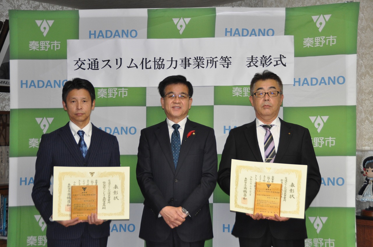 両手を組んで立っている高橋市長の両側に盾の様な物と賞状を持った男性が立ち、記念撮影をしている写真