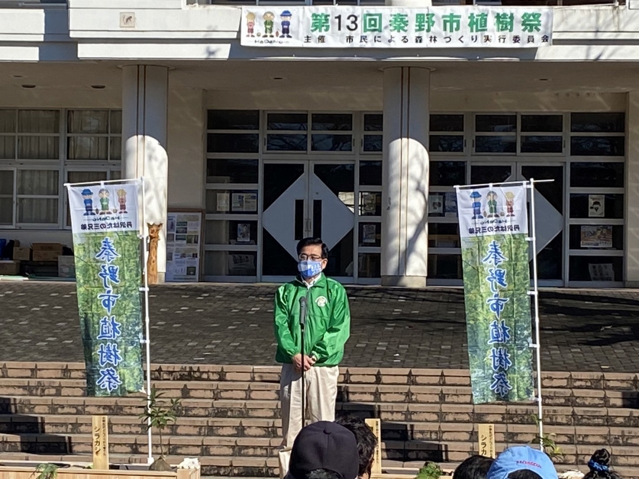 入り口上部に「第13回秦野市植樹祭」の横断幕が掲げられている建物の前で、緑色のジャケットを着た高橋市長がマイクスタンドの前に立ち話をしている写真