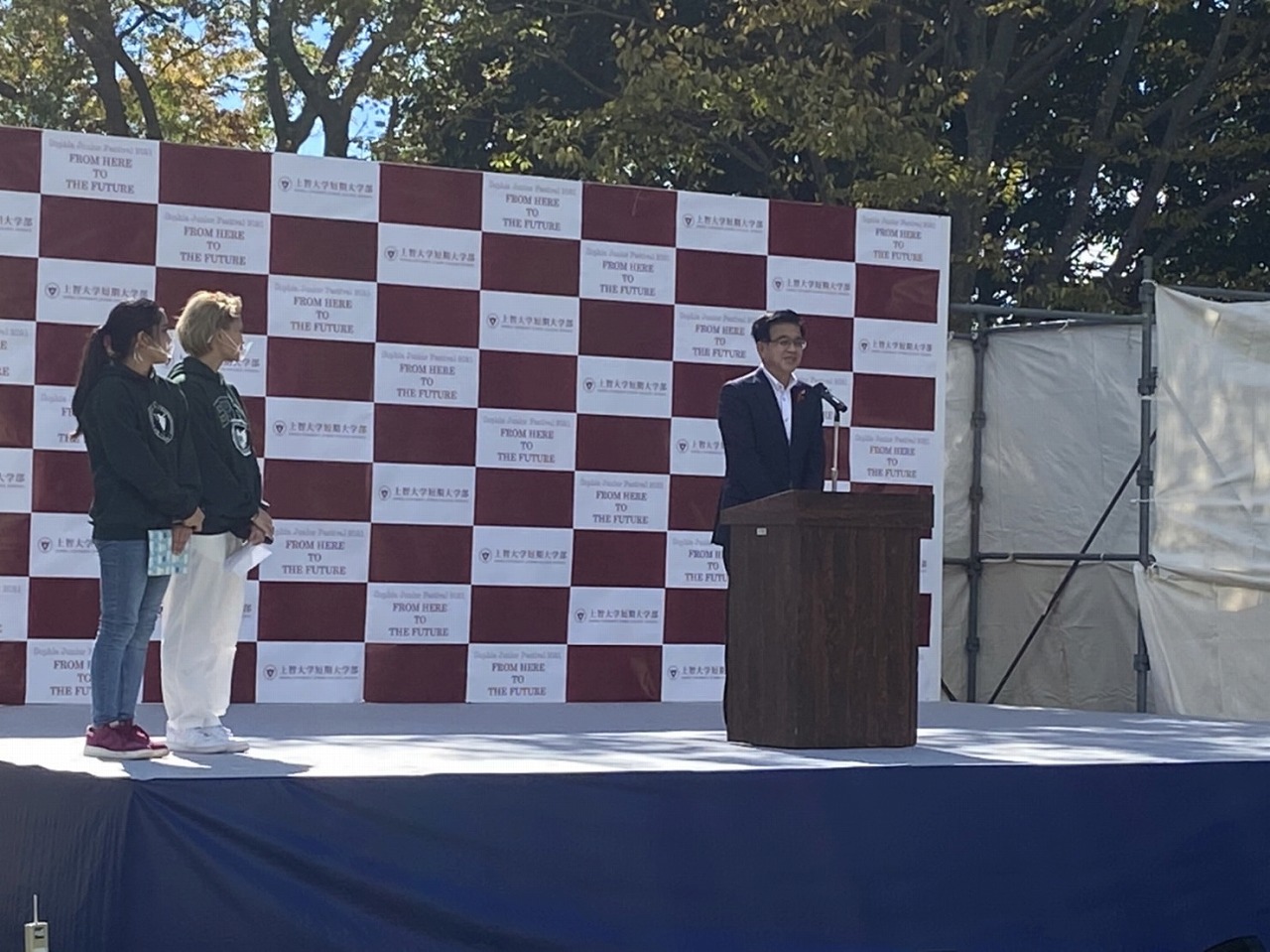 赤と白の市松模様のバックパネルが設置された屋外ステージ上で、高橋市長が演台に立ち話をしている写真