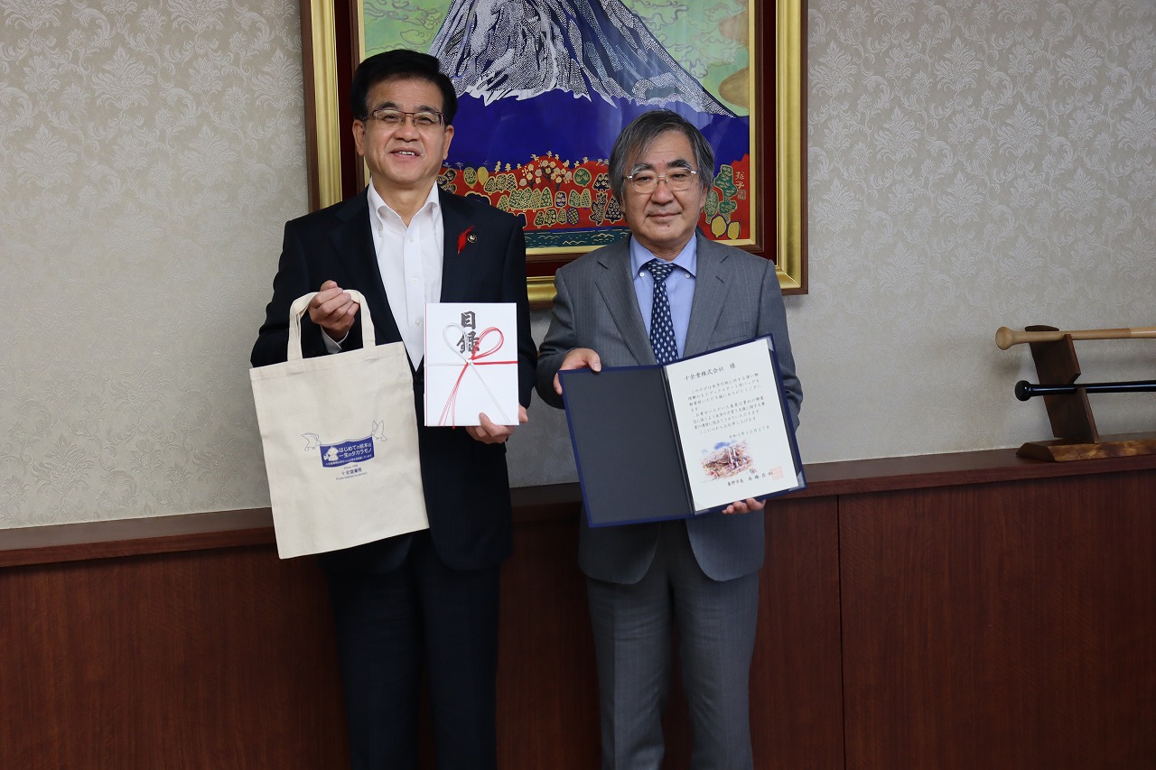 コットンバックと目録を持っている高橋市長と証書ホルダーを開いて持っている大島代表取締役社長が並んで記念撮影をしている写真