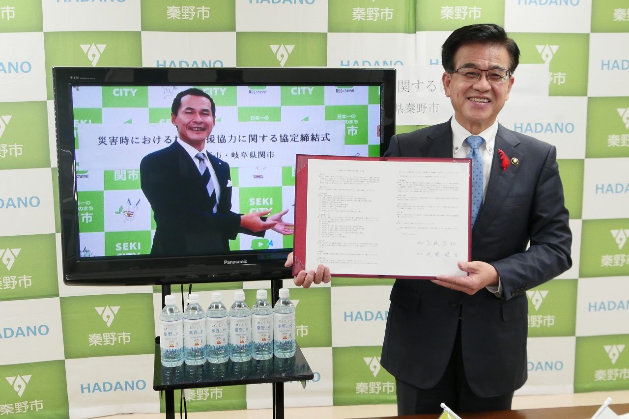 カメラに笑顔を向ける高橋市長が、協定書を手に持ち、モニターに映し出された別の男性とともに秦野市と災害時における相互協力協定を締結している様子を写した写真