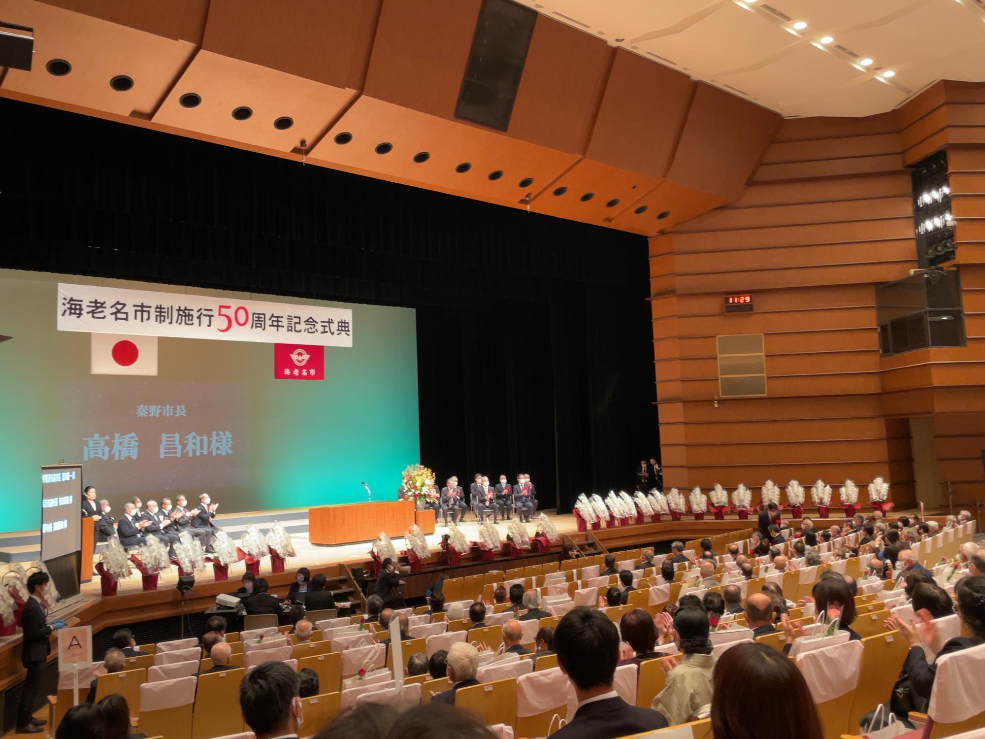 「海老名市制施行50周年記念式典」と書かれた横断幕が掲げられたコンサートホールのような会場のステージ上に、大勢の人が登壇し、聴衆が多数着席している海老名市市制施行50周年記念式典の様子写真