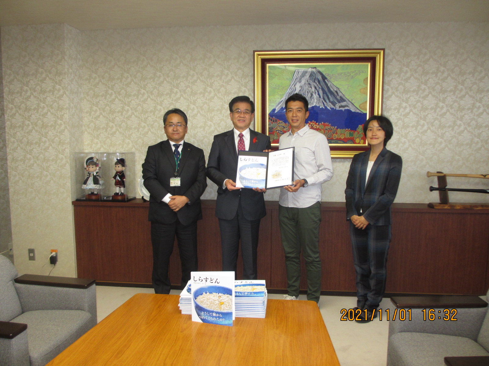 スーツやシャツ姿の4人の男女が並んで立ち、中央の高橋市長と男性が冊子のようなものを一緒に持ち記念撮影をしている写真