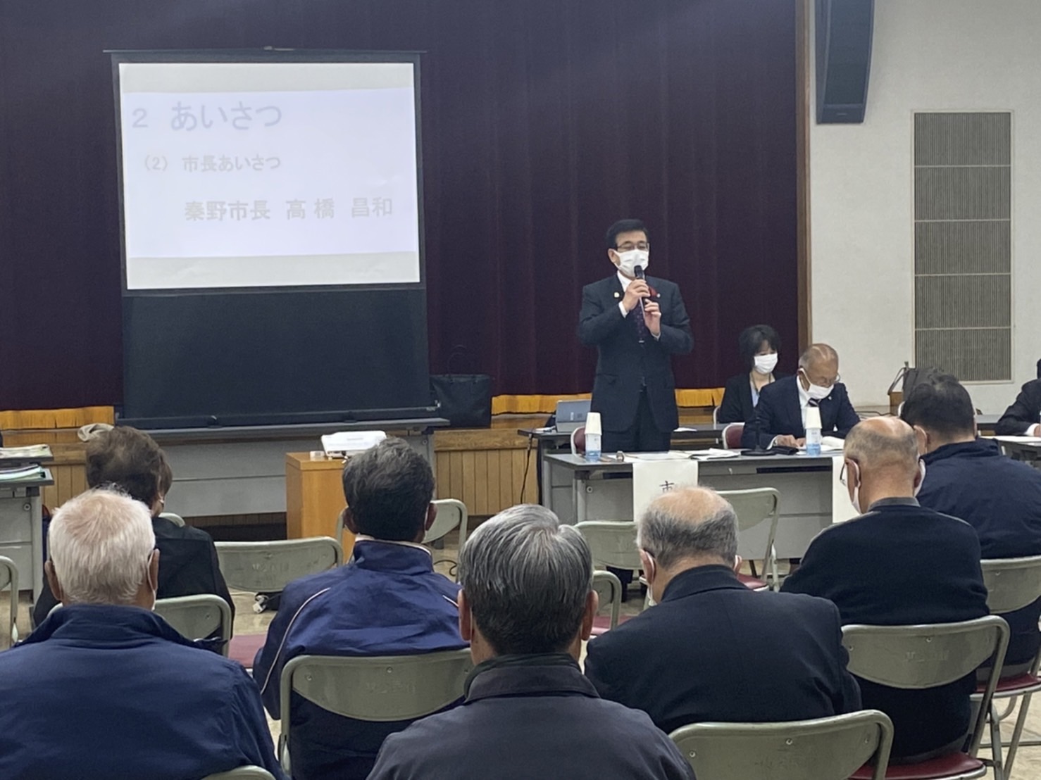 「2 あいさつ 市長あいさつ 秦野市長 高橋 昌和」の文字が映し出されたスクリーンの前で、スーツ姿の高橋市長がマイクを持って聴衆に向かって話をしている様子の写真