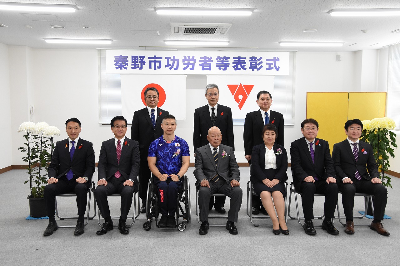 「秦野市功労者等表彰式」と書かれた横断幕が掲げられた室内で、車椅子の男性を含む複数の人々が並んで座り、その後ろに別の男性たちが立っている集合写真