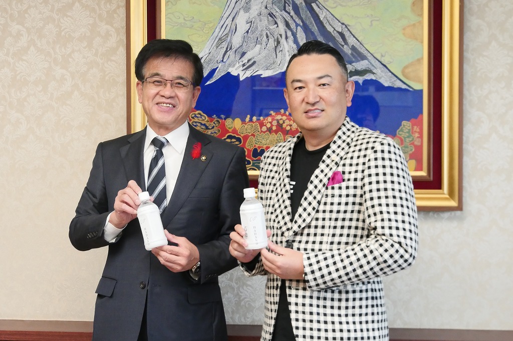 スーツ姿の高橋市長とチェック柄のジャケットを着た岩田代表の二人が、白いボトルを手に笑顔で記念撮影を行っている写真