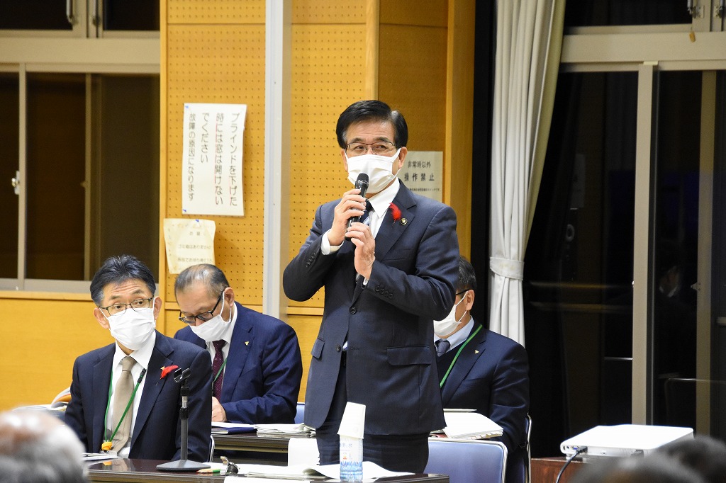 周囲のテーブルに他のスーツ姿の男性が着席している中、スーツ姿の高橋市長が両手でマイクを持ち発言している様子を捉えた写真