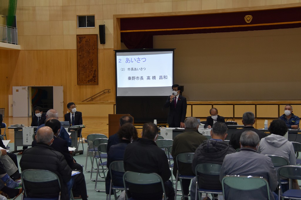 体育館のような場所で、スクリーンに「あいさつ」と映し出される中、スーツ姿の高橋市長が聴衆に向かってマイクで話している様子を捉えた写真