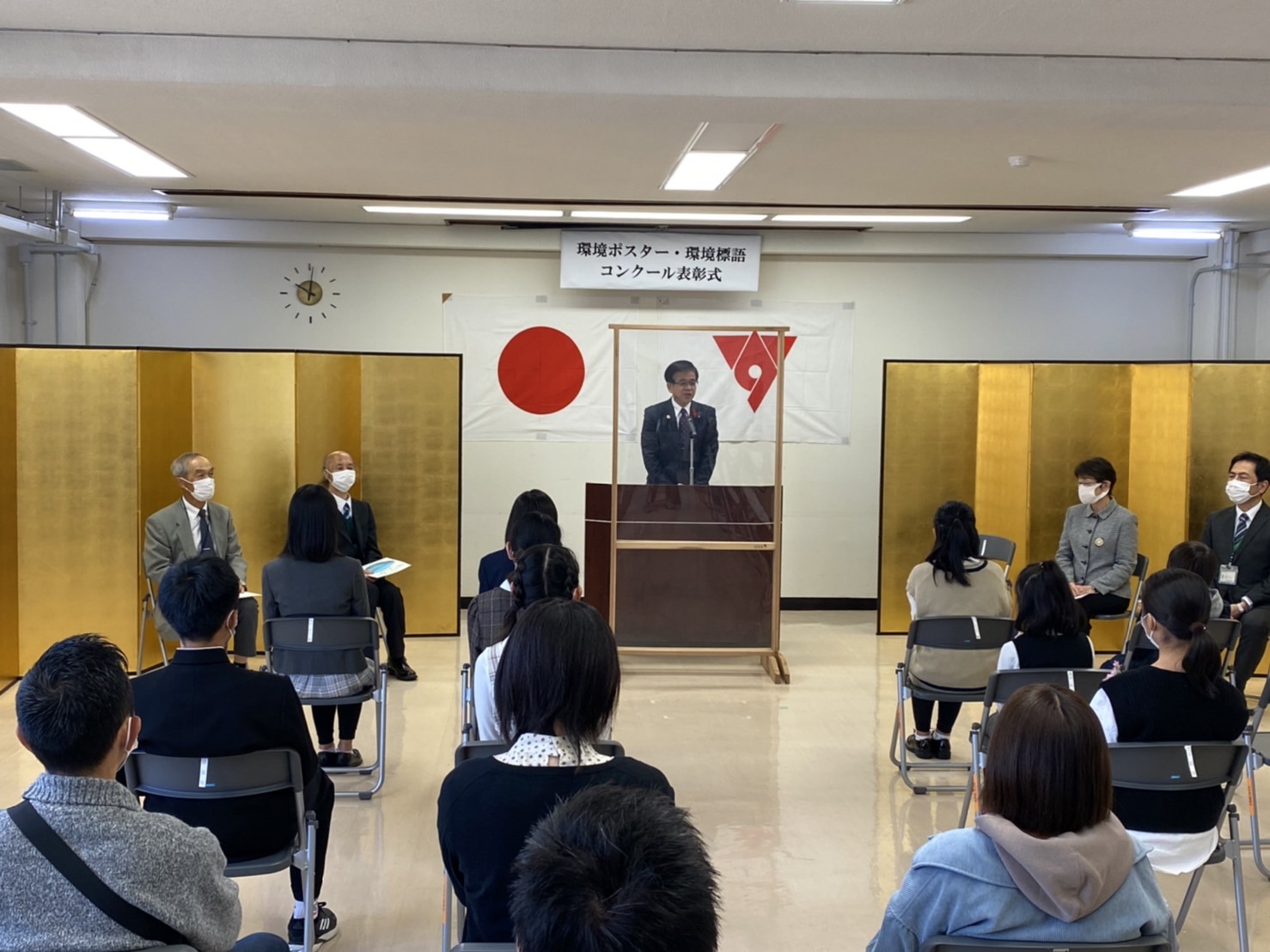 「環境ポスター・環境標語コンクール表彰式」と書かれた横断幕と日の丸の旗、市章が掲げられ、金屏風が設置された室内で、クリアボードが設置された演台に立ち話をしている高橋市長の写真