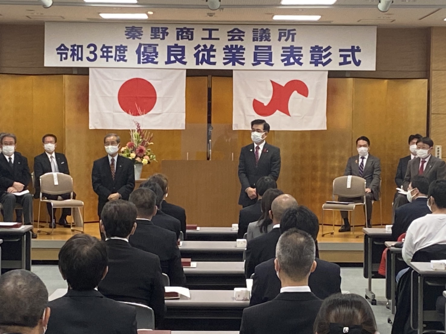 「秦野商工会議所 令和3年度 優良従業員表彰式」と書かれた横断幕と日の丸の旗が掲げられ、金屏風が設置された会場で、スーツ姿の出席者たちが整然と座る前に立っている高橋市長の写真