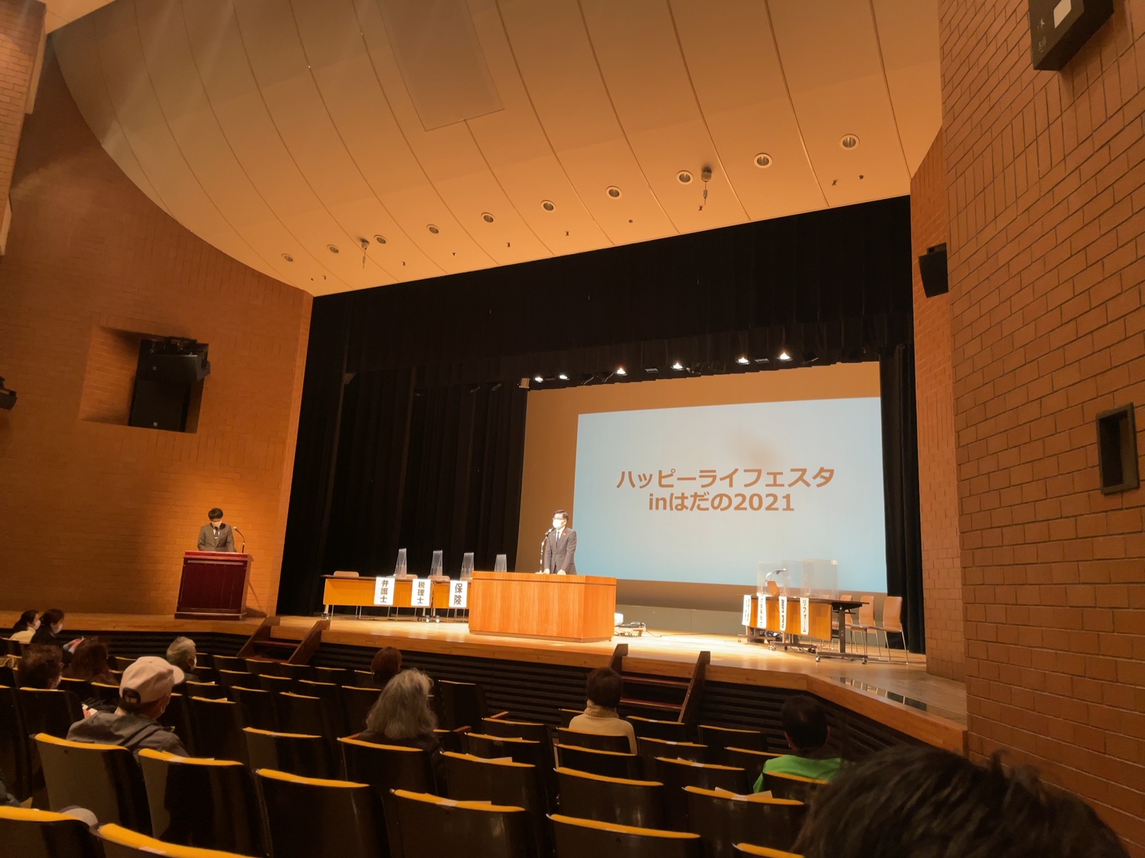 「ハッピーライフフェスタ inはだの2021」と書かれたスクリーンが設置されている広々とした講堂の舞台で、マスクを着用した高橋市長が演台の前に立ち、聴衆に向かって話をしている様子の写真