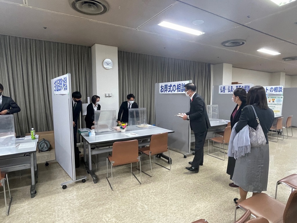 複数のブースがパーティションで仕切られた室内で、高橋市長が「お葬式の相談」と書かれているブースの前に立ち、相談員と思われる人に話しかけている様子の写真