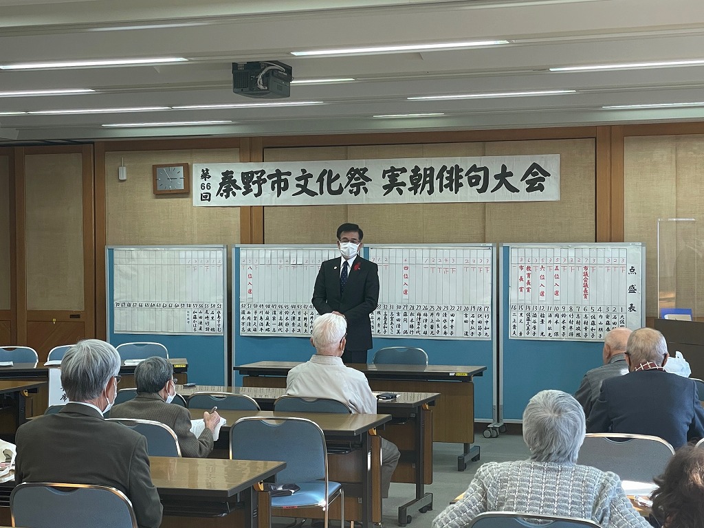 「第66回秦野市文化祭 実朝俳句大会」と書かれた紙が貼られた室内前方で、スーツ姿でマスクを着けた高橋市長が、多数の参加者が着席する前でマイクを使わずに話している写真