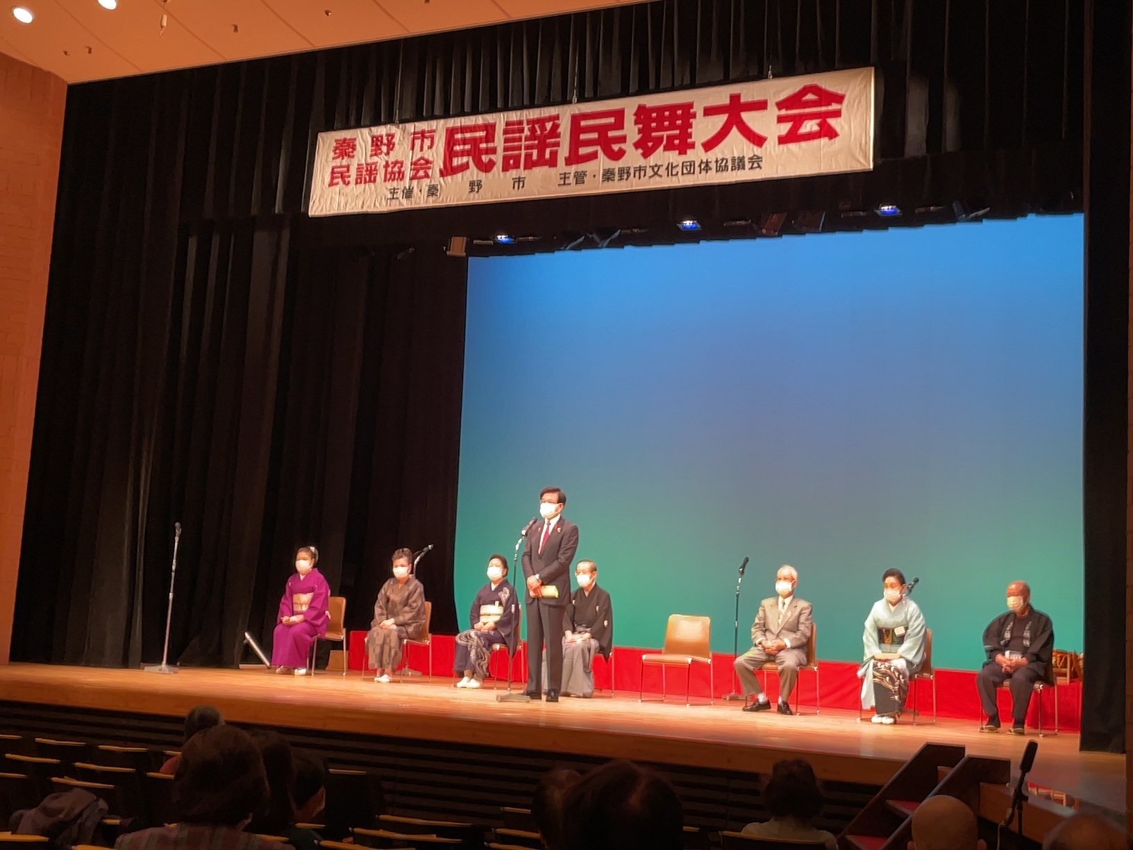 「秦野市民謡協会 民謡民舞大会」と書かれた横断幕が掲げられたステージ上で、和装やスーツ姿の男女7人が間隔を空けて椅子に座る中、中央でスーツに赤いネクタイを締めた高橋市長がマイクの前に立ち、観客に向かって挨拶をしている様子の写真