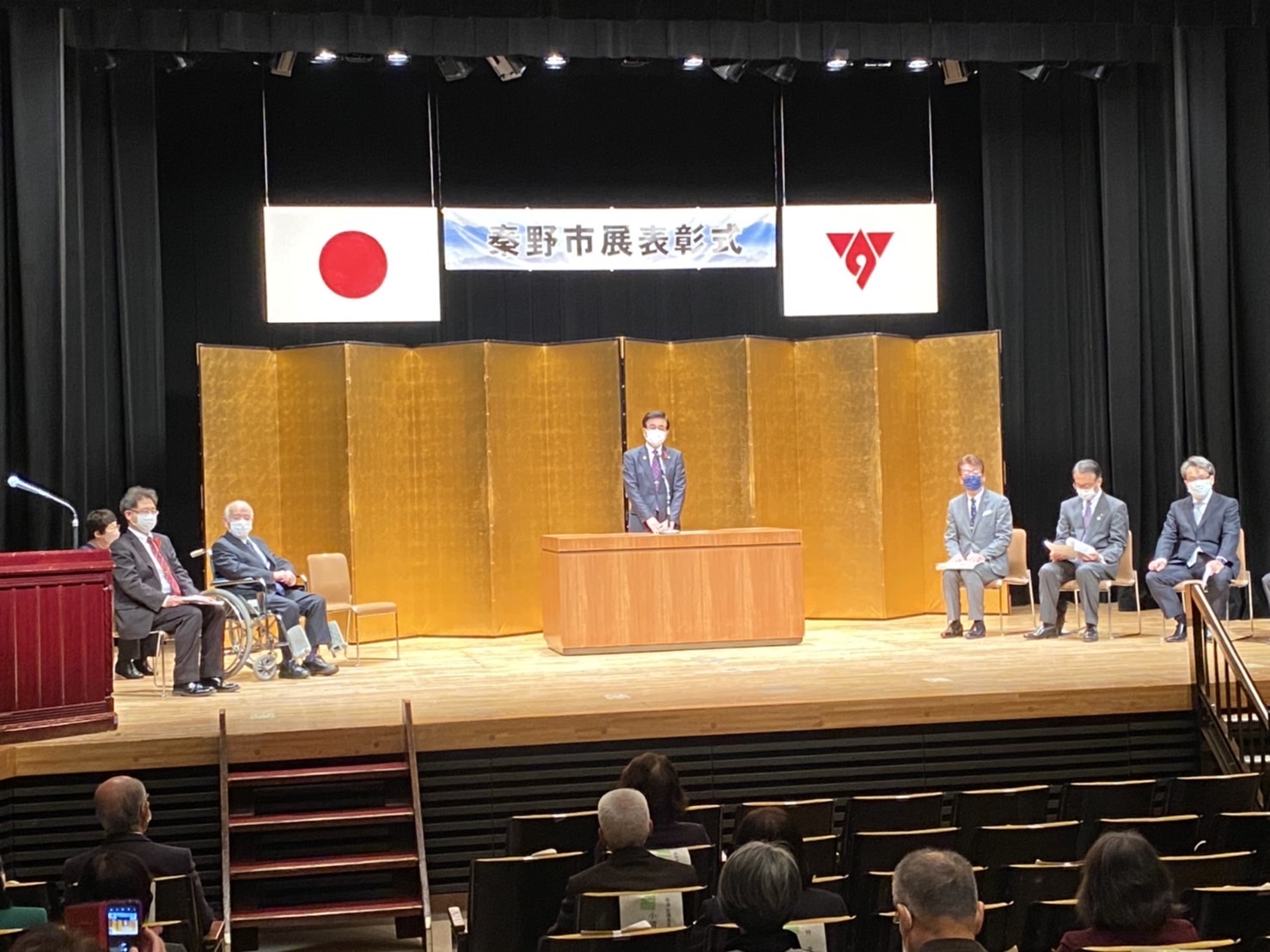 「秦野市展表彰式」と書かれた横断幕と金屏風が設置されているステージ上でスーツ姿の複数の男性が椅子に座り、中央の演台に高橋市長が立ち話をしている様子の写真