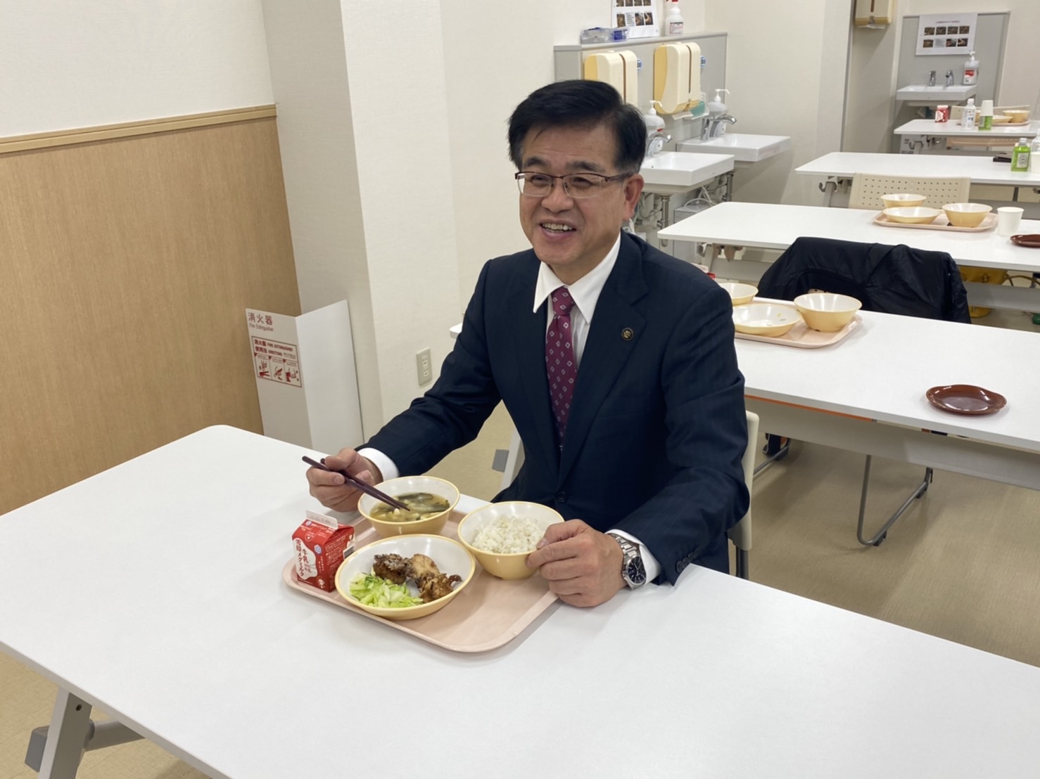 スーツ姿の高橋市長が箸を手に笑顔で給食のトレイを前に座り、給食を試食している様子の写真