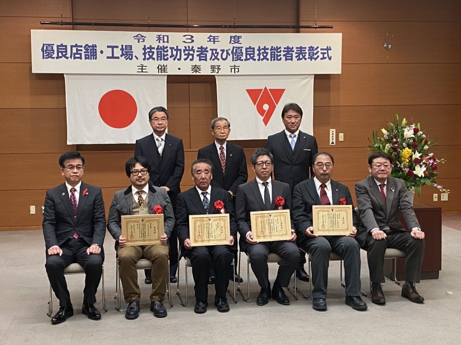 複数のスーツ姿の男性が、横断幕と日の丸の旗を背景に記念撮影しており、中央に座っている数名が表彰状のようなものを持っている優良店舗・工場及び技能者表彰式での集合写真