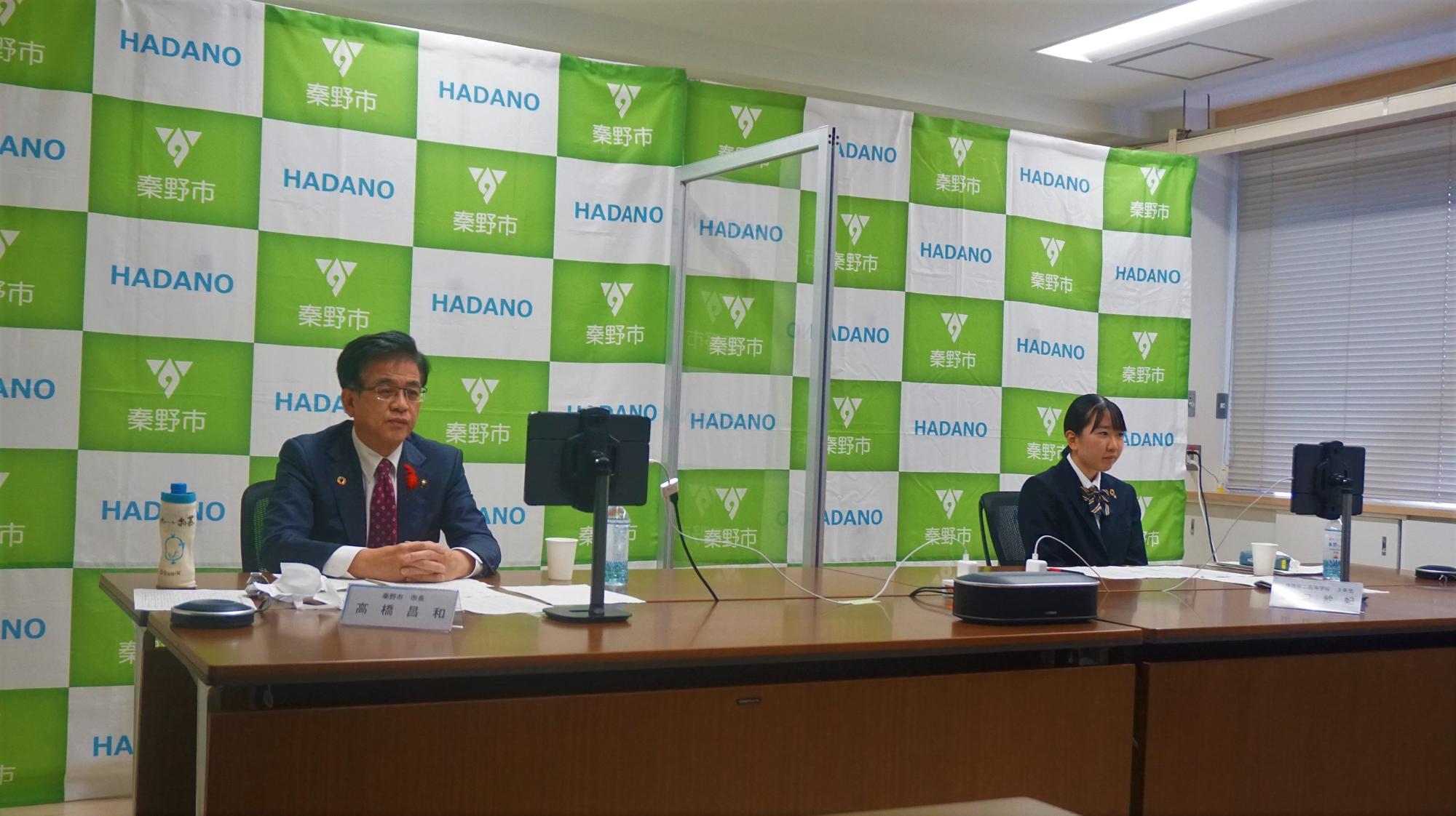 秦野市のバックパネルを背景に、高橋市長と制服姿の女子生徒が横並びに並んで座り全国報徳サミット掛川市大会が行われている様子を写した写真