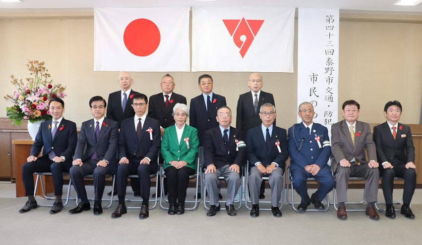 垂れ幕と日の丸、秦野市の市章が掲げられている室内で複数のスーツ姿の男性と中央に座る緑色のジャケットを着た女性が椅子に座り記念撮影を行っている写真