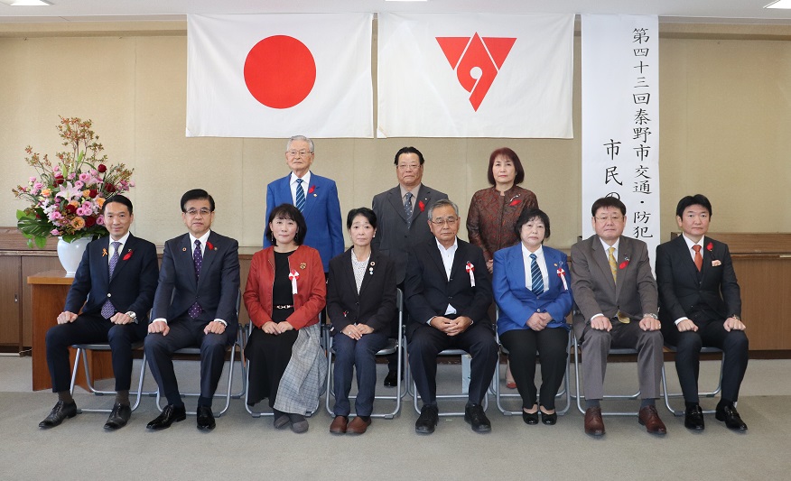 椅子に座る複数の男女が、背景の日の丸の旗と秦野市の市章、白い垂れ幕を背に並び集合写真を撮っている写真