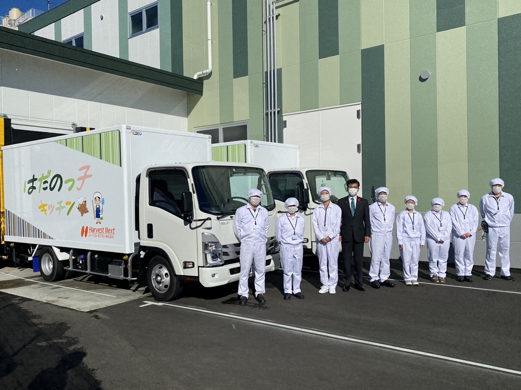 新しい建物と「はだのっ子キッチン」と書かれた給食配送車を背景に、高橋市長と白衣と帽子を身につけたスタッフたちが並んで立っている様子を捉えた写真