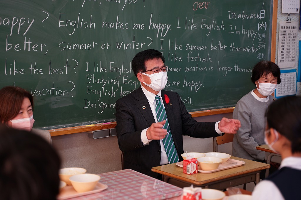 黒板に書かれた英語の文章を背景に、高橋市長が給食の入ったトレーの前に座り、両手を広げて生徒たちに何かを熱心に話している中学校での一コマを捉えた写真