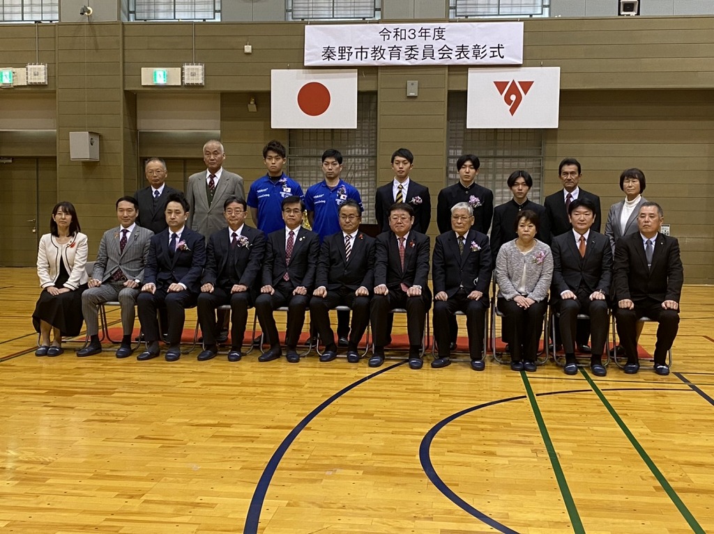 屋内スポーツ施設の木製の床の上で、秦野市教育委員会表彰式の横断幕を背景に、複数のスーツ姿の大人たちが集まり、記念撮影をしている写真