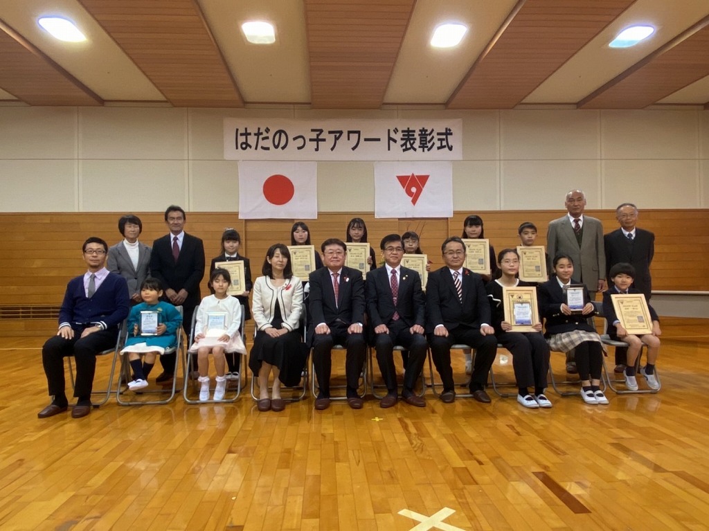 木製の床の上で「はだのっ子アワード表彰式」と書かれた横断幕を背景に、スーツ姿の大人たちと子供たちが表彰状のようなものを持って並んでいる記念写真