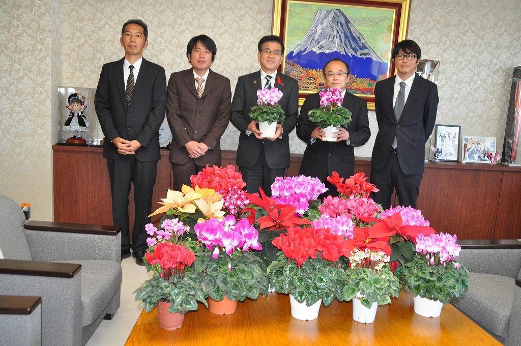 たくさんのカラフルなシクラメンの鉢植えが並べられたテーブルを前に、高橋市長を中心にスーツ姿の4名の男性が並んで立ち、カメラに笑顔を向け記念撮影を行っている写真