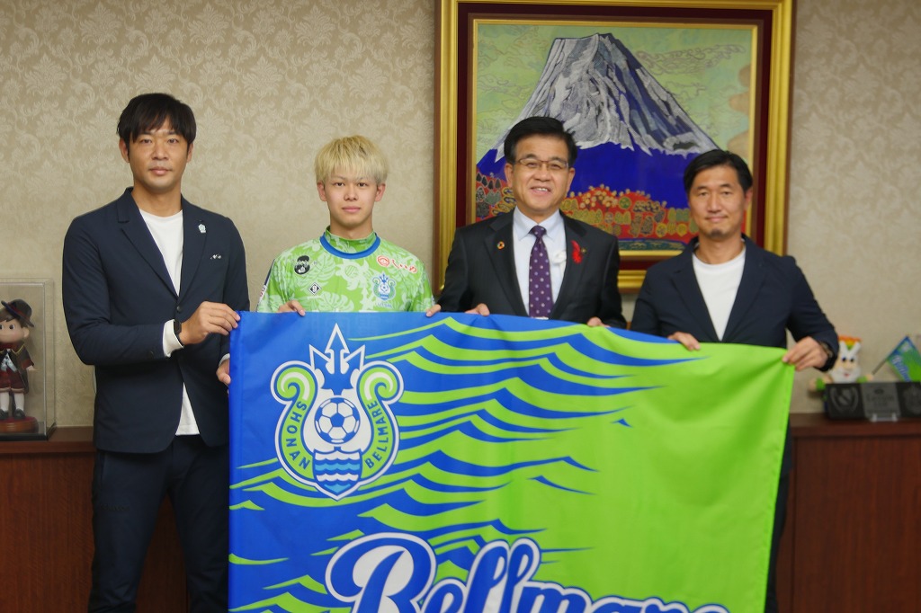 中央に薮内選手と高橋市長、その両脇に立つ2名の男性とともに青と緑のフラッグを持ち記念撮影をしている写真