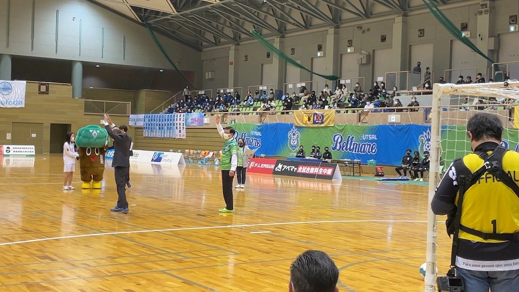 体育館で行われたフットサルの試合のイベントで、マスコットキャラクターのもりりんとスーツ姿の男性が、観客席に向かって手を振っている様子を遠景で捉えた写真