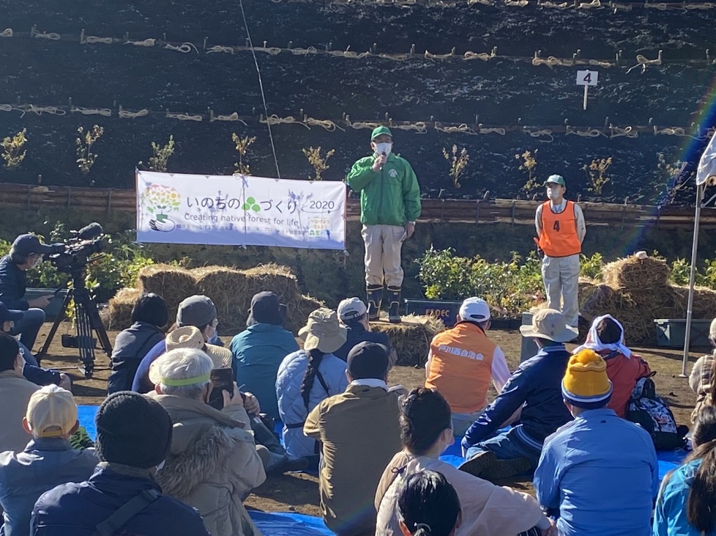 大勢の人が地面に座り、マイクを片手に持った男性が「いのちの森づくり」と書かれた横断幕の前で話している、新東名秦野丹沢植樹祭2021のイベントの様子を撮影した写真