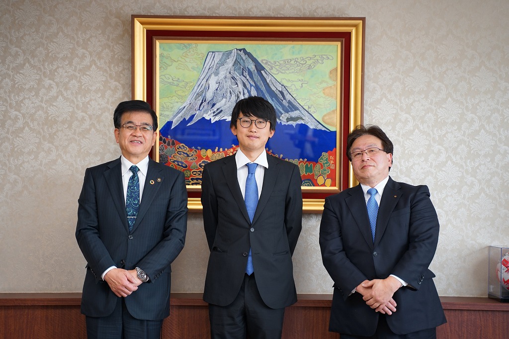 山の絵画が飾られた壁の前で、高橋市長、須知代表取締役、関係者の男性が並んで立ち、記念撮影をしている写真