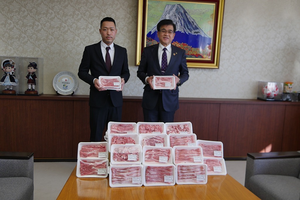 多くの豚肉のパックが積み重ねられたテーブルを前にして、高橋市長と門倉敏夫さんが豚肉のパックを持ってカメラに向かって立ち、記念撮影をしている写真